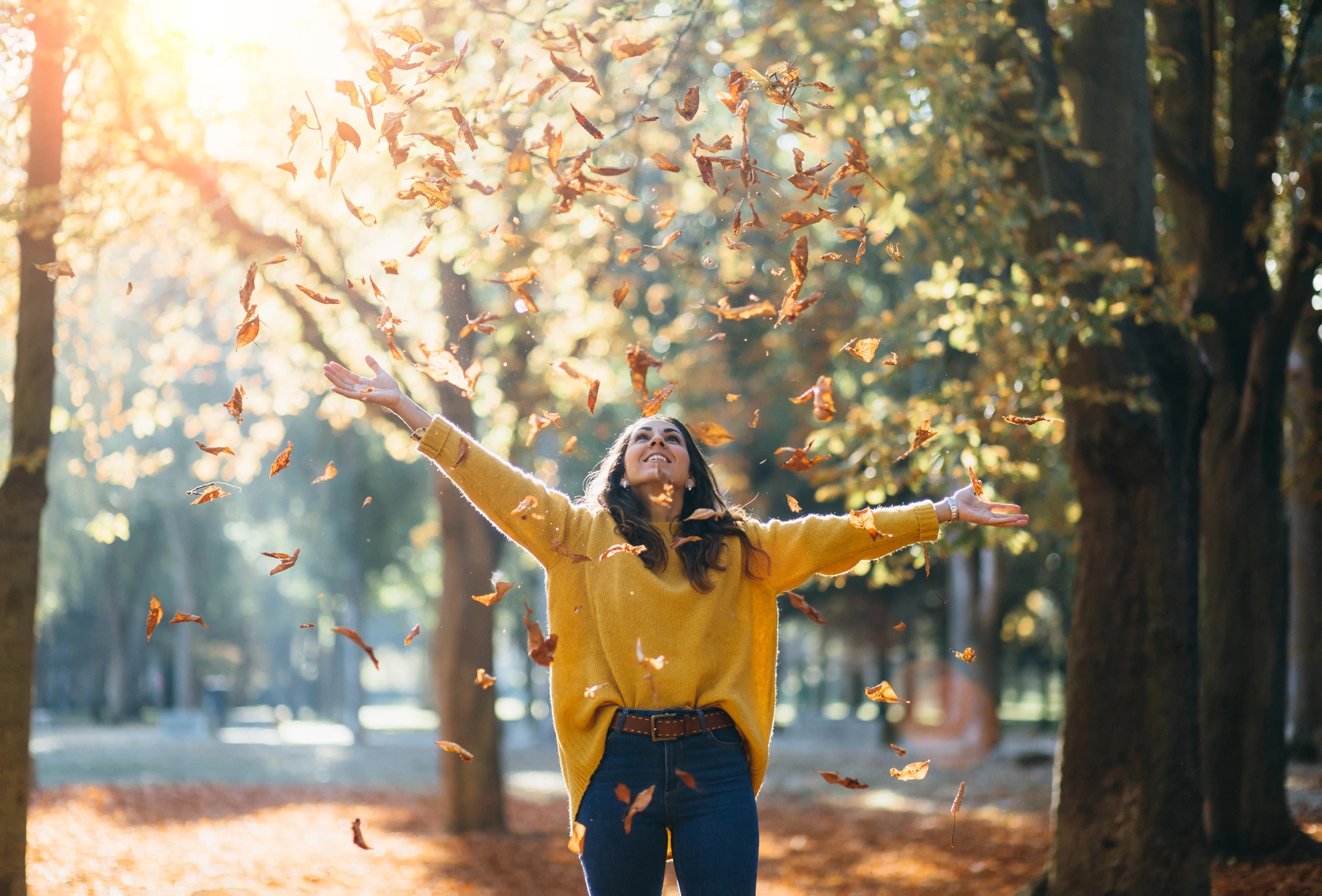 Jak dbać o zdrowie jesienią? Odporność na jesień. Kobieta w żółtym swetrze podrzuca do góry jesienne liście na spacerze w parku.
