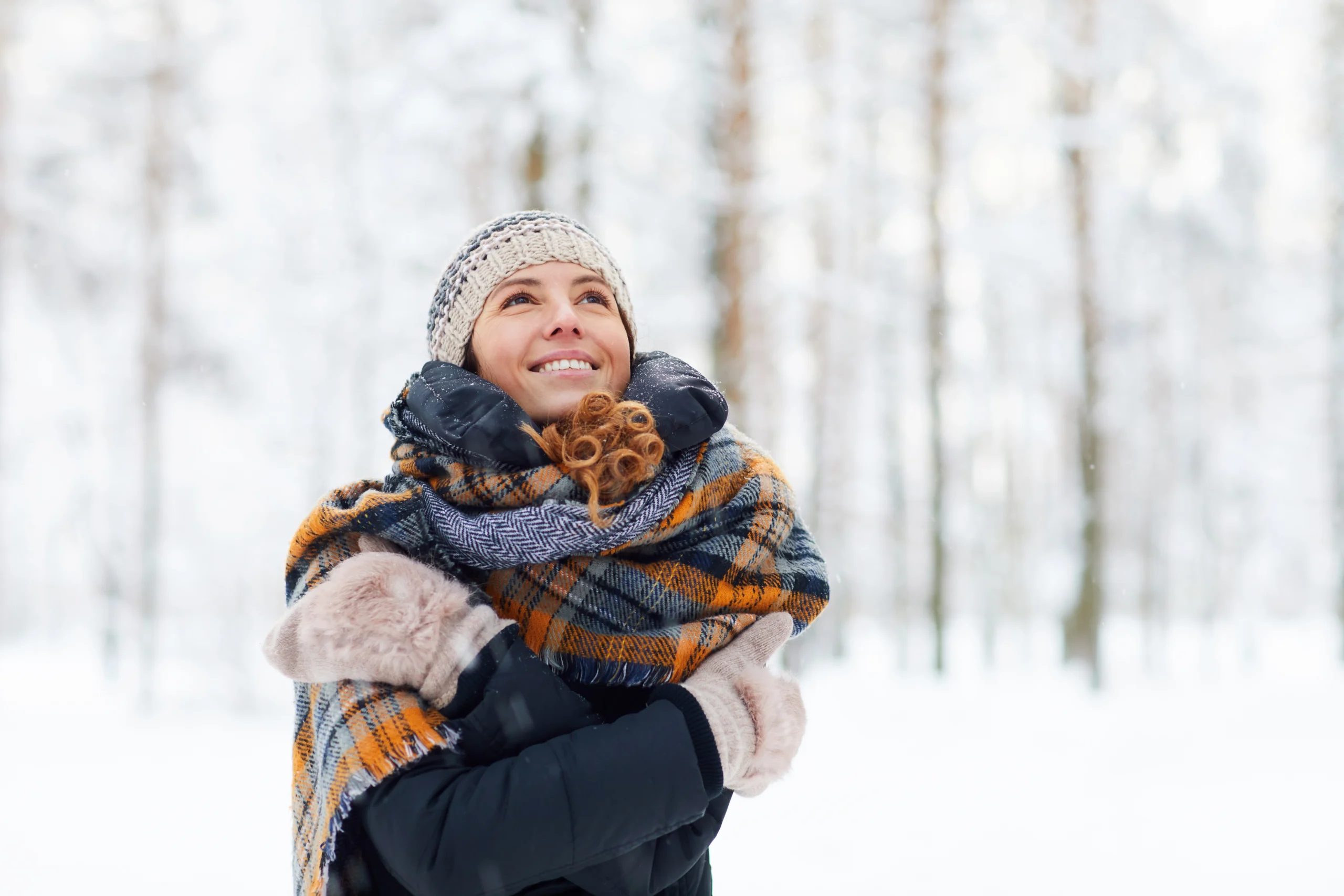 Naturalne sposoby na odporność. Kobieta w czapce i rękawiczkach otula się szalikiem, stojąc w lesie na śniegu.