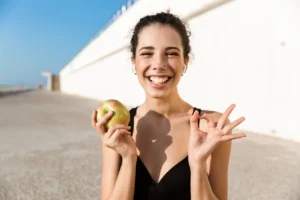 Jakie suplementy na tarczycę mogą Ci pomóc utrzymać ją w dobrej kondycji? Jak zadbać o zdrowie tarczycy? Uśmiechnięta kobieta trzyma jabłko w dłoni i cieszy się słoneczną pogodą.