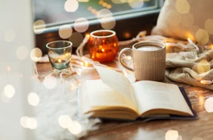 Hygge, duńska filozofia życia, pozwala odnaleźć radość i spokój nawet w długie jesienne czy zimowe wieczory. Zbliżenie na otwartą książkę, kubek kakao i świeczki, leżące na parapecie w zimowy dzień.