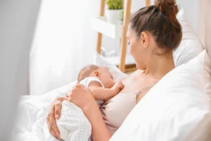 Młoda mama karmi piersią niemowlaka, siedząc na kanapie w domu, opierając się o białą poduszkę.