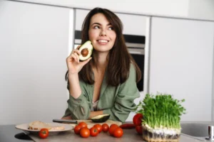 Uśmiechnięta kobieta w zielonej koszuli, o pięknej cerze i grubych, gęstych, ciemnych włosach, opiera się o blat kuchenny, trzymając w ręku połówkę awokado.