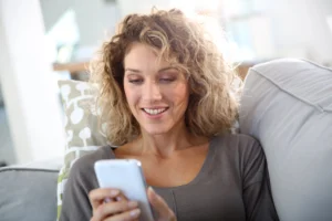 Kobieta o blond kręconych włosach, w szarej bluzce, siedzi na kanapie i sprawdza na telefonie jakie właściwości ma fitochinon, czyli witamina K1.