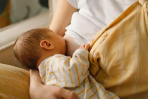 Młoda mama, ubrana w biały t-shirt i żółtą koszulę, karmi piersią niemowlę.